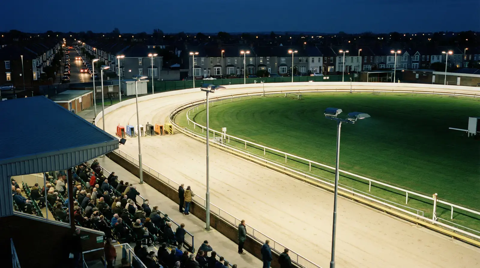 Aerial view of a UK greyhound racing track at night with floodlights illuminating the sand oval and six traps